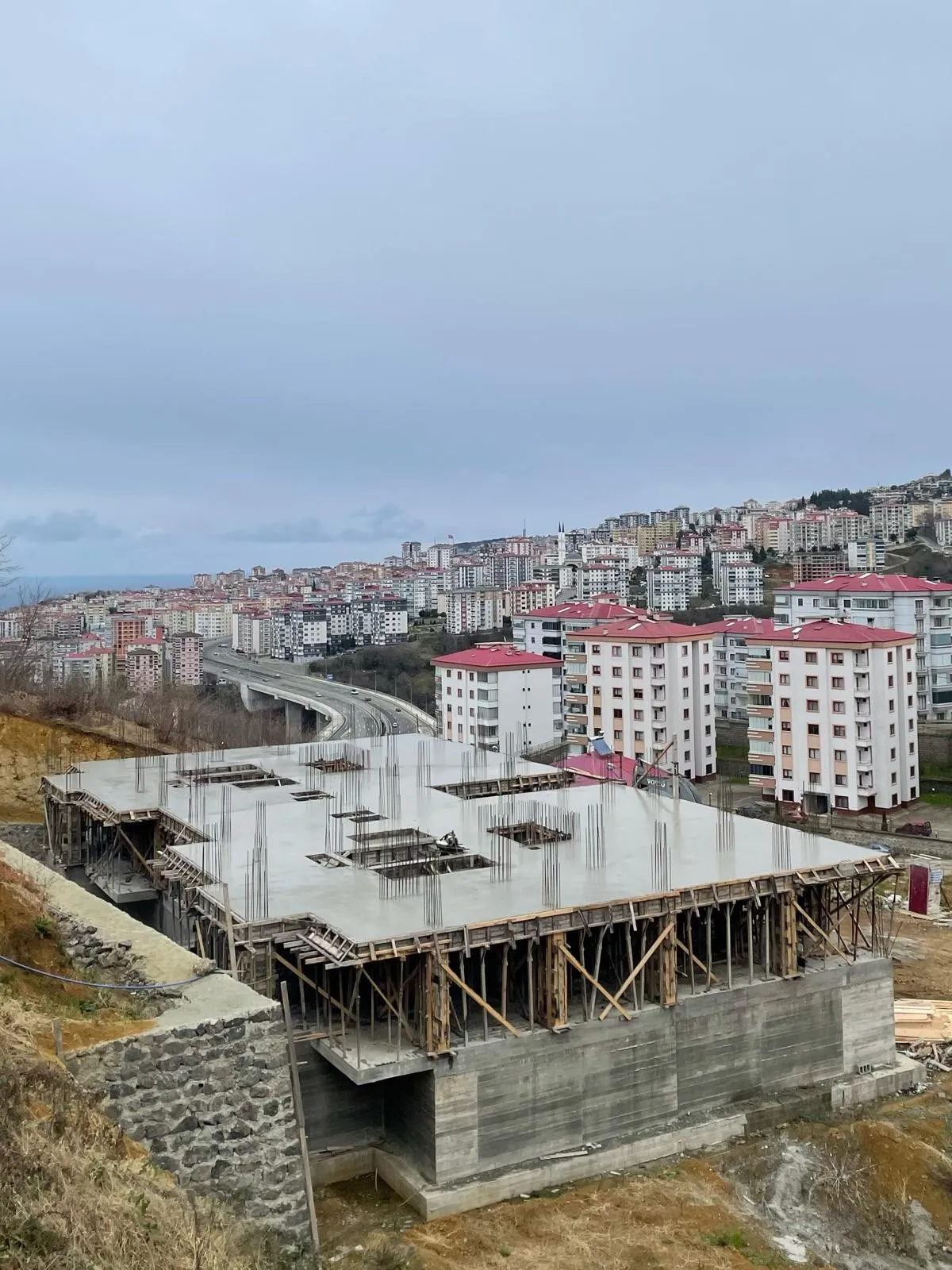 Yamaçta dökülmüş beton döşeme, filizleri çıkmış kolon demirleri, arkada şehir ve viyadük